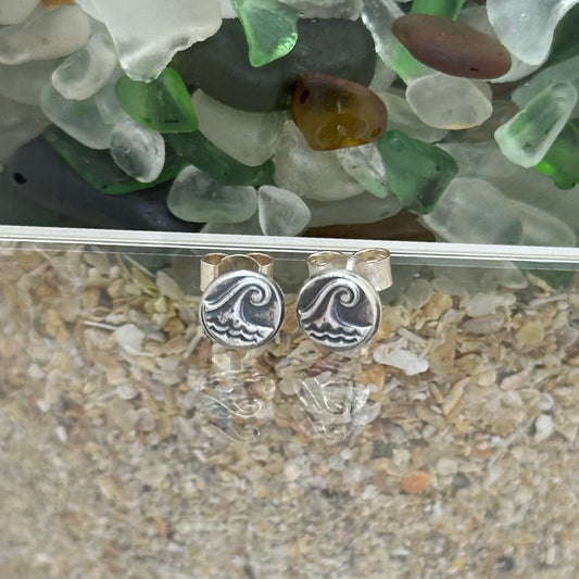 Handmade sterling silver pebble stud earrings with an embossed wave design and light patina, displayed against a coastal background.