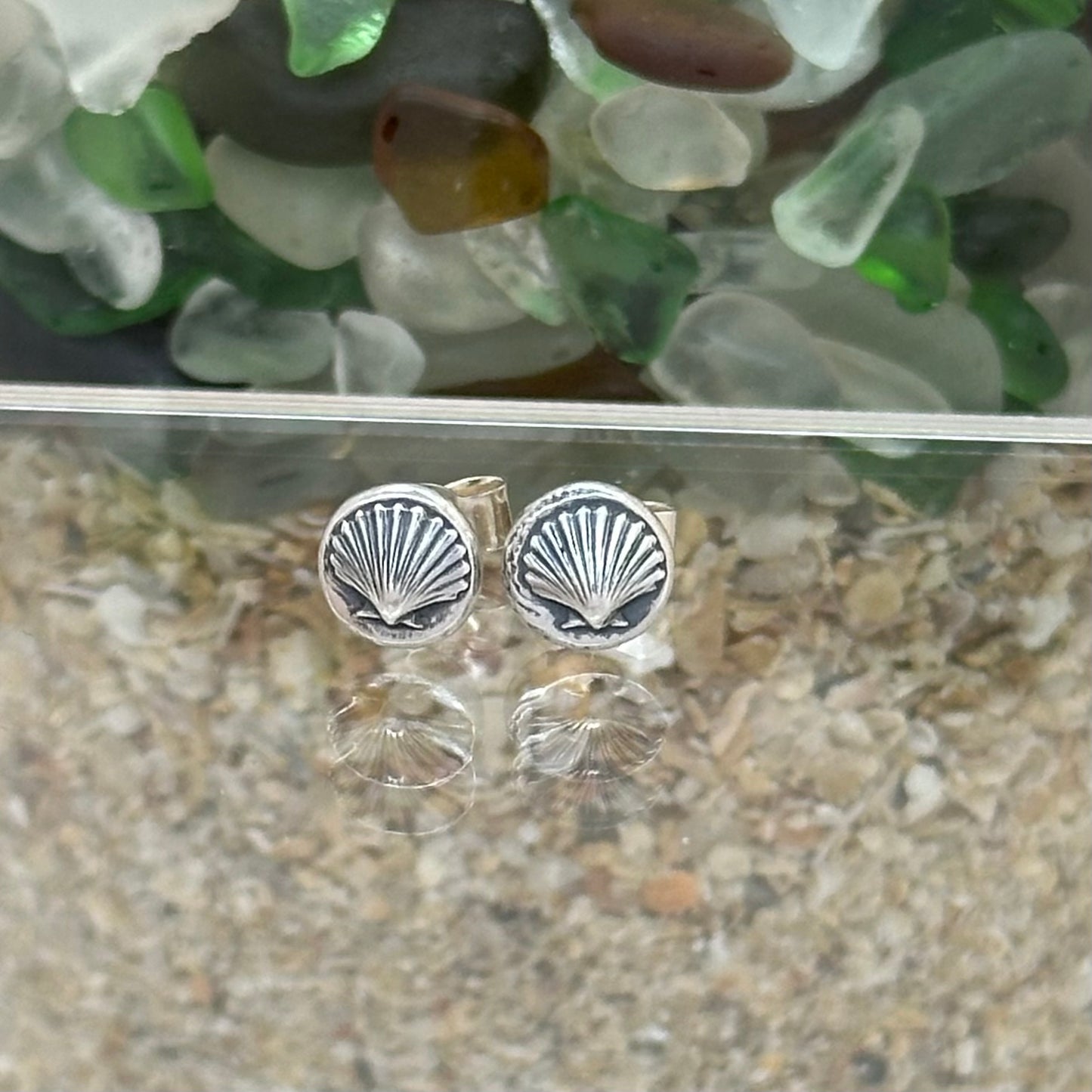 Handmade sterling silver pebble stud earrings with an embossed shell design and light patina, displayed against a coastal background