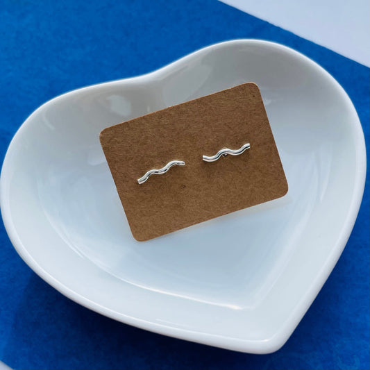 A pair of sterling silver squiggle bar stud earrings displayed on a recycled kraft card, placed inside a white heart-shaped dish on a blue background. The earrings feature a smooth, wavy design with a polished finish