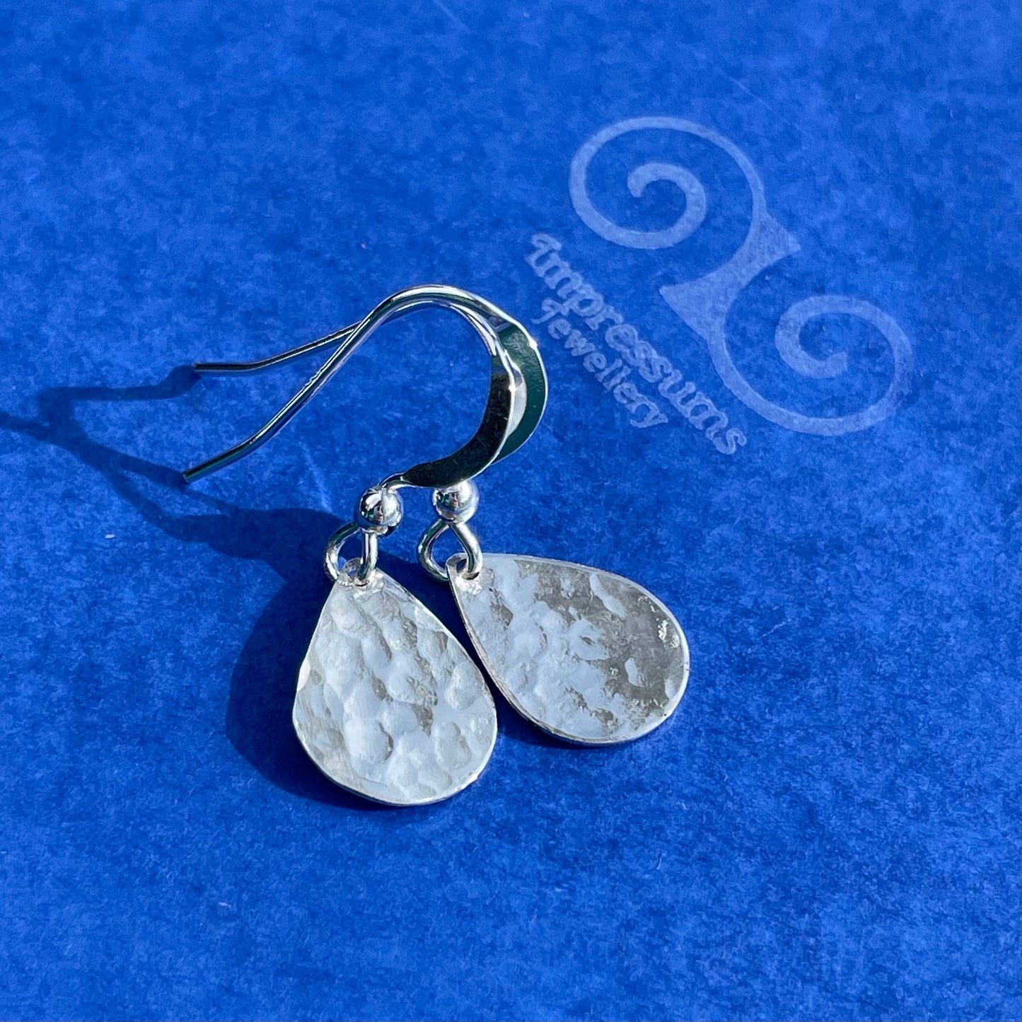 Handmade sterling silver teardrop dangle earrings with a hammered texture, displayed against a dark blue background.